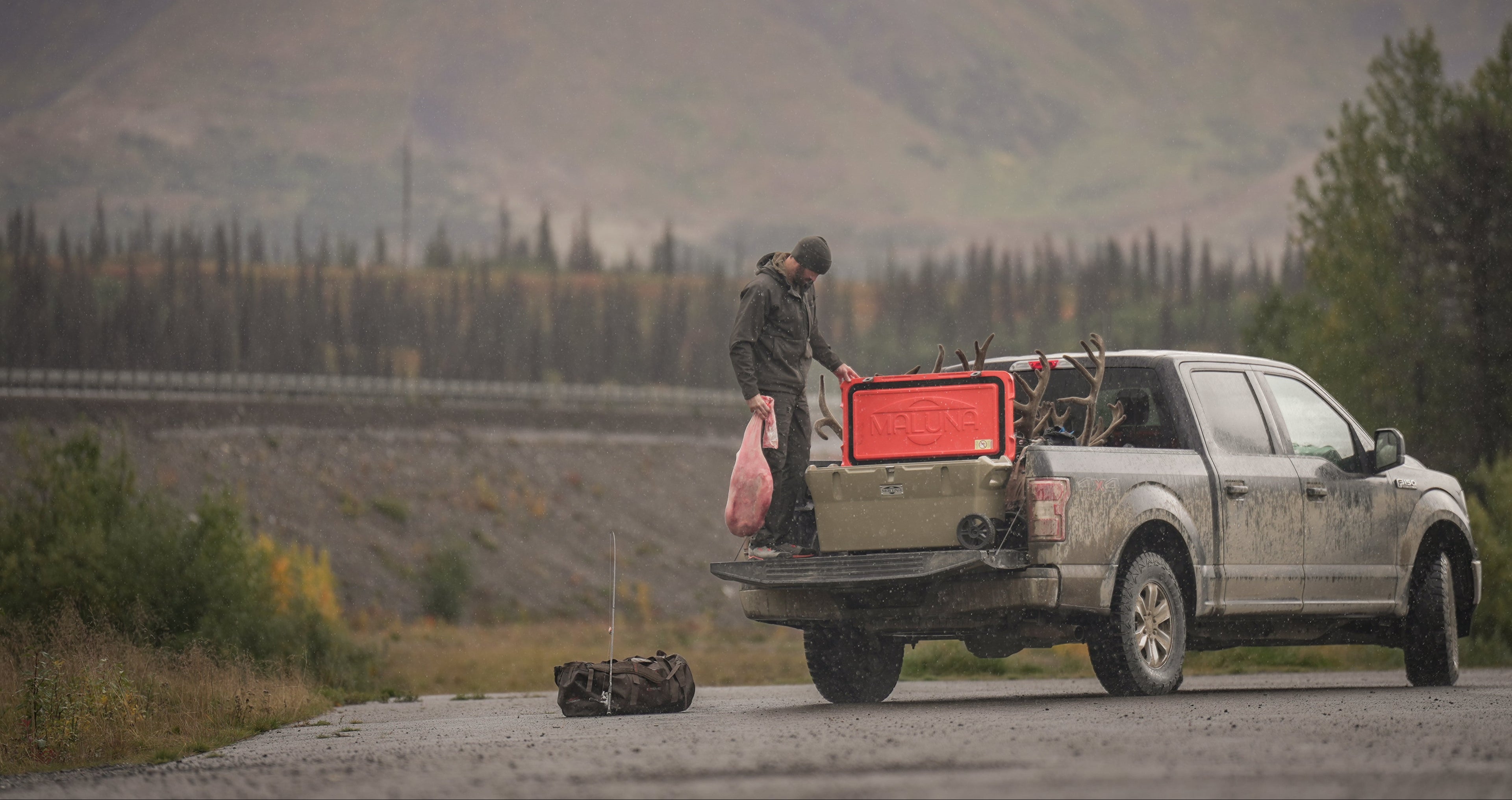 A person stands on the open tailgate of a dusty pickup truck on a road, handling a pink bag of game near a cooler in an outdoor mountain and forest setting.