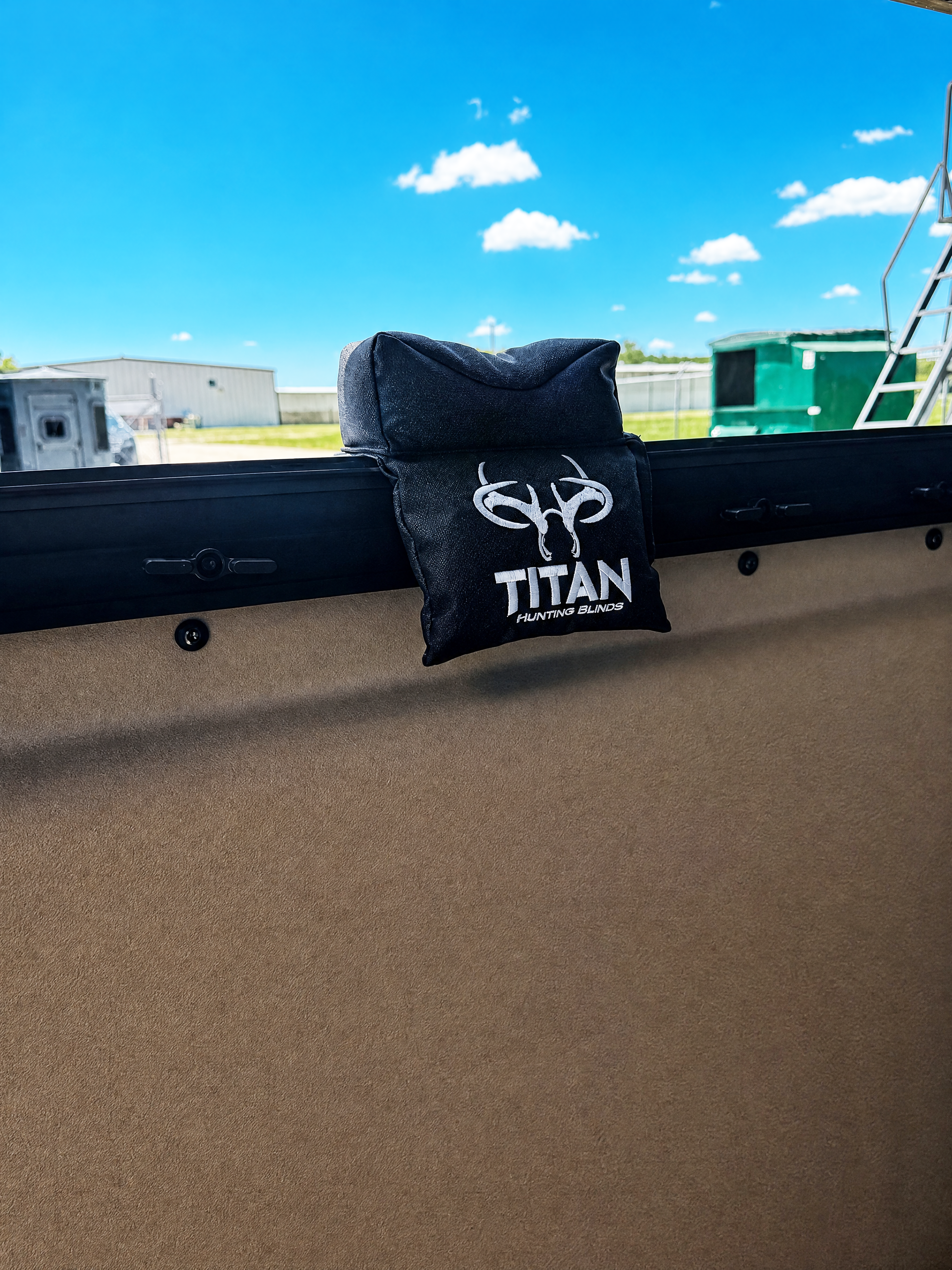 A black fabric window shooting rest bag with a white printed logo and the word 'TITAN' hanging from a black metal frame against an outdoor backdrop with a blue sky and clouds.