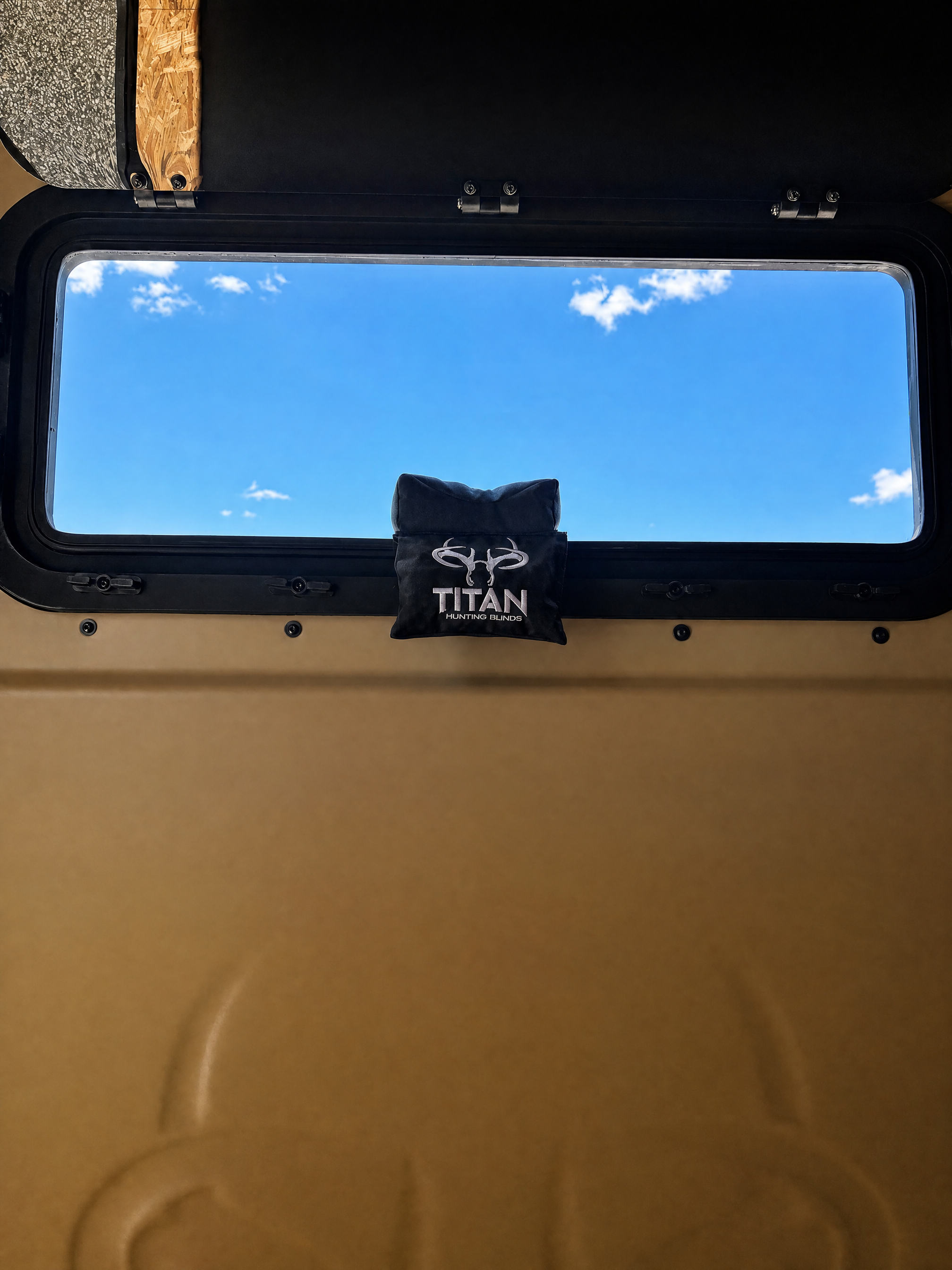 A black fabric window shooting rest bag with a white printed logo and the word 'TITAN' hanging from a black metal frame against an outdoor backdrop with a blue sky and clouds.