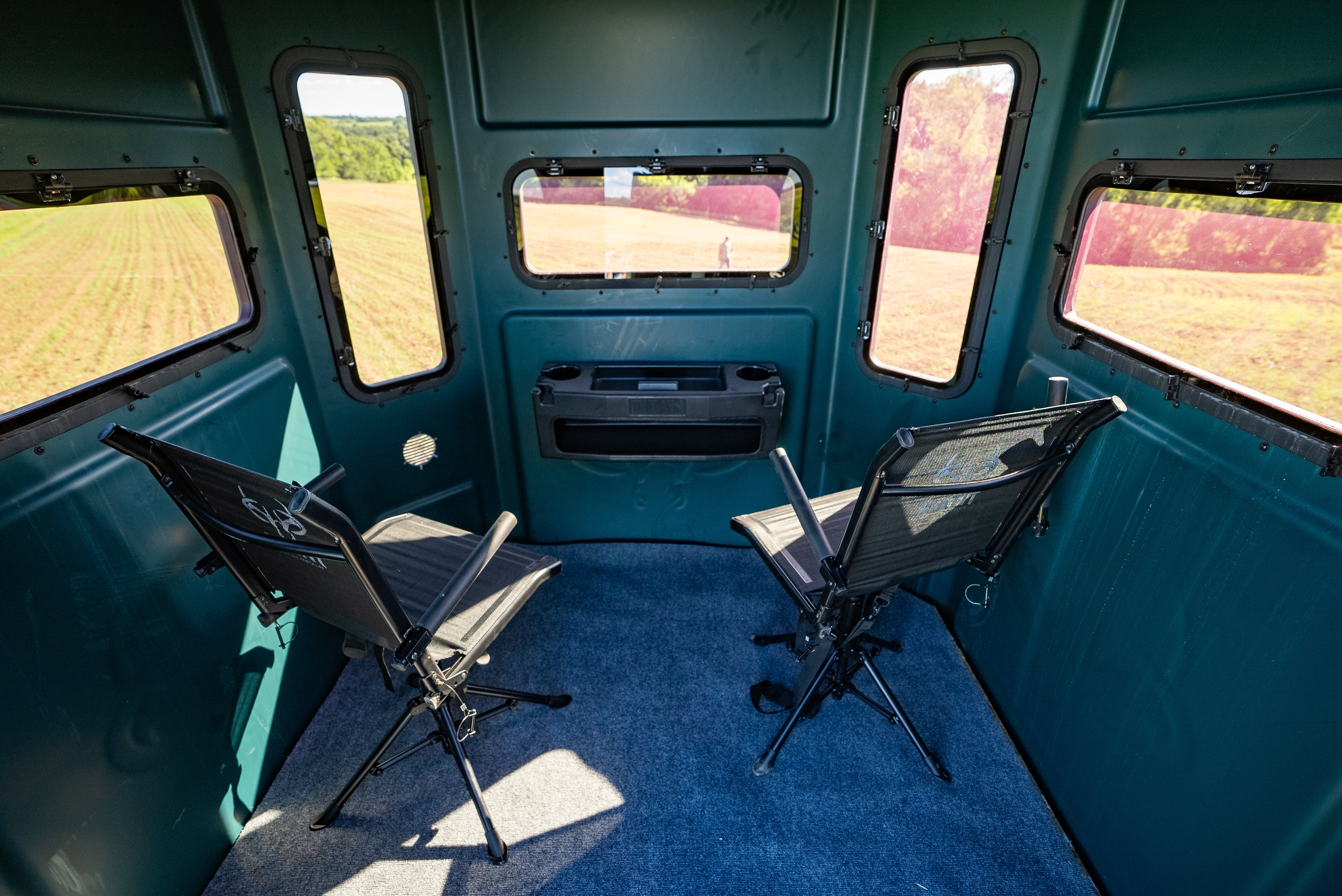 The interior of a teal hunting blind with two black mesh chairs facing a central window, set against a background of open field visible through the windows.