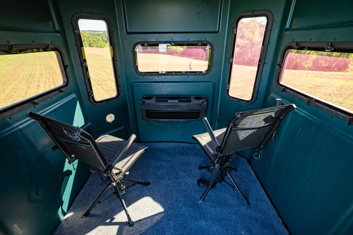 The interior of a teal hunting blind with two black mesh chairs facing a central window, set against a background of open field visible through the windows.
